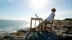 Man with a laptop sitting in chair at wooden desk on a beach